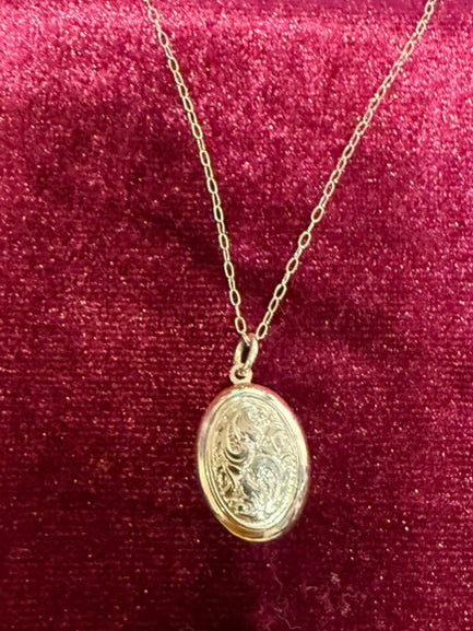 A gold oval pendant necklace with an engraved religious design, displayed on a burgundy velvet jewelry cushion over a dark gray keyboard.